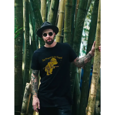 Man wearing a black 'Get Clapped' t-shirt featuring a playful Winnie the Pooh design, standing amidst bamboo.